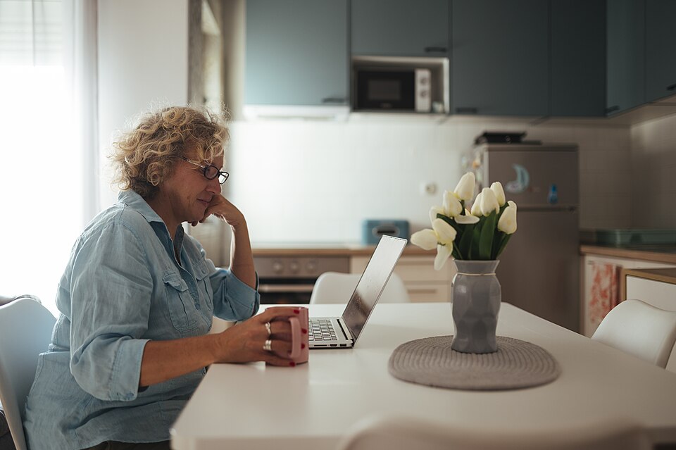 A woman working on her laptop at a bright kitchen table with flowers and coffee
